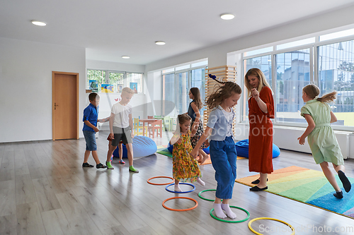 Image of Small nursery school children with female teacher on floor indoors in classroom, doing exercise. Jumping over hula hoop circles track on the floor.