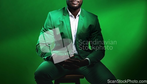 Image of Fashion, suit and black man model with a formal outfit sitting on a chair while posing in a studio. Elegant, trendy and African male in luxury, stylish and fancy clothes isolated by green background.