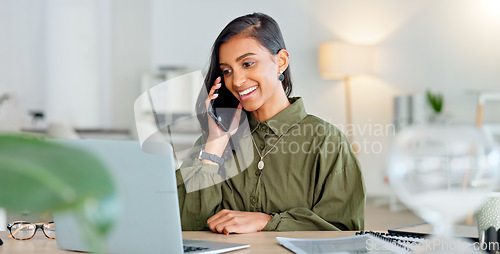 Image of Happy business woman talking on phone call or young entrepreneur answering cellphone while sitting in front of work laptop in an office. Indian female executive smiling and laughing at a funny joke.