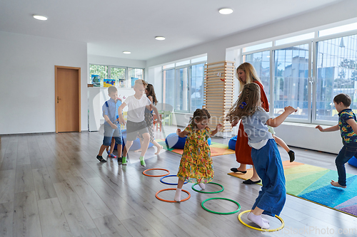 Image of Small nursery school children with female teacher on floor indoors in classroom, doing exercise. Jumping over hula hoop circles track on the floor.