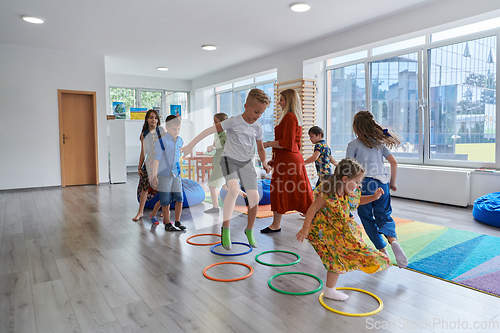 Image of Small nursery school children with female teacher on floor indoors in classroom, doing exercise. Jumping over hula hoop circles track on the floor.