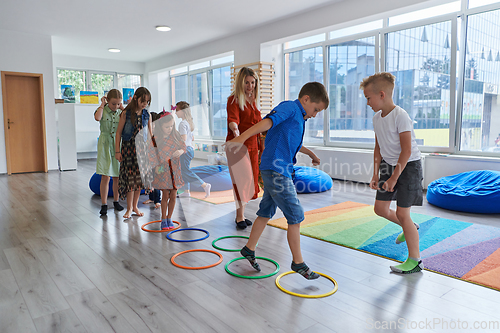 Image of Small nursery school children with female teacher on floor indoors in classroom, doing exercise. Jumping over hula hoop circles track on the floor.