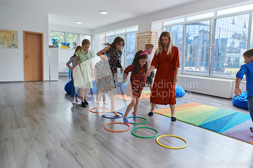 Image of Small nursery school children with female teacher on floor indoors in classroom, doing exercise. Jumping over hula hoop circles track on the floor.