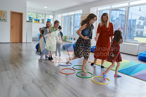 Image of Small nursery school children with female teacher on floor indoors in classroom, doing exercise. Jumping over hula hoop circles track on the floor.