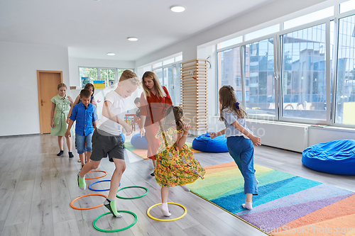 Image of Small nursery school children with female teacher on floor indoors in classroom, doing exercise. Jumping over hula hoop circles track on the floor.