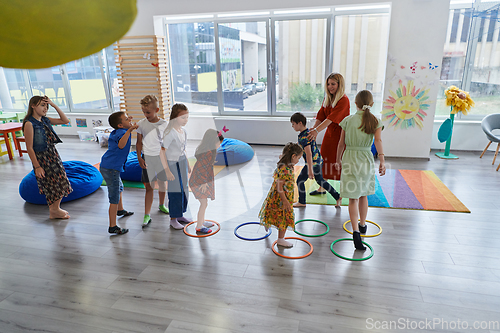 Image of Small nursery school children with female teacher on floor indoors in classroom, doing exercise. Jumping over hula hoop circles track on the floor.