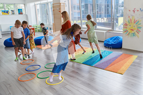 Image of Small nursery school children with female teacher on floor indoors in classroom, doing exercise. Jumping over hula hoop circles track on the floor.