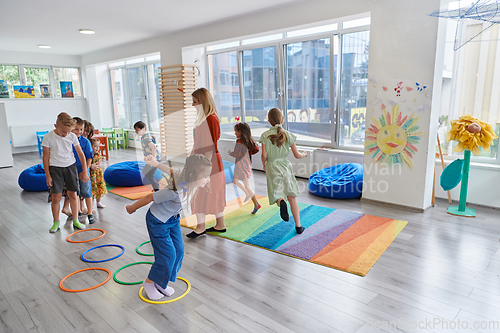 Image of Small nursery school children with female teacher on floor indoors in classroom, doing exercise. Jumping over hula hoop circles track on the floor.