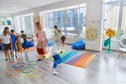 Image of Small nursery school children with female teacher on floor indoors in classroom, doing exercise. Jumping over hula hoop circles track on the floor.
