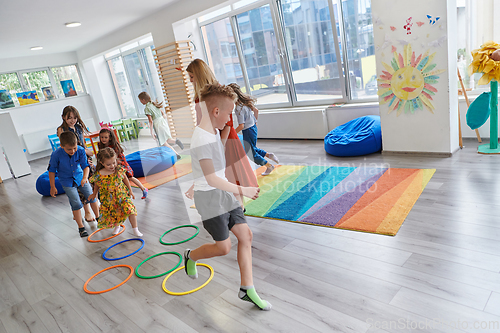 Image of Small nursery school children with female teacher on floor indoors in classroom, doing exercise. Jumping over hula hoop circles track on the floor.