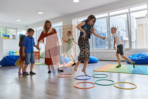 Image of Small nursery school children with female teacher on floor indoors in classroom, doing exercise. Jumping over hula hoop circles track on the floor.