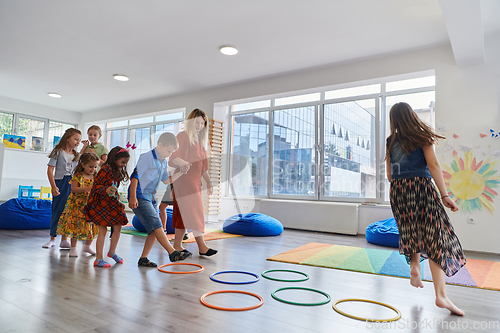 Image of Small nursery school children with female teacher on floor indoors in classroom, doing exercise. Jumping over hula hoop circles track on the floor.