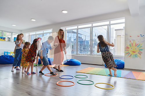 Image of Small nursery school children with female teacher on floor indoors in classroom, doing exercise. Jumping over hula hoop circles track on the floor.