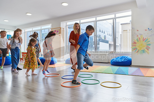 Image of Small nursery school children with female teacher on floor indoors in classroom, doing exercise. Jumping over hula hoop circles track on the floor.