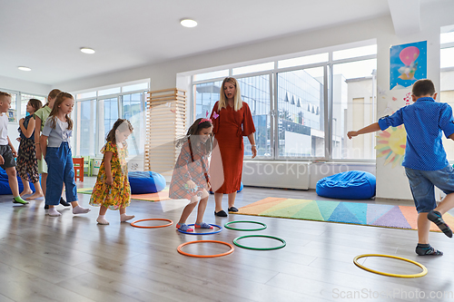 Image of Small nursery school children with female teacher on floor indoors in classroom, doing exercise. Jumping over hula hoop circles track on the floor.