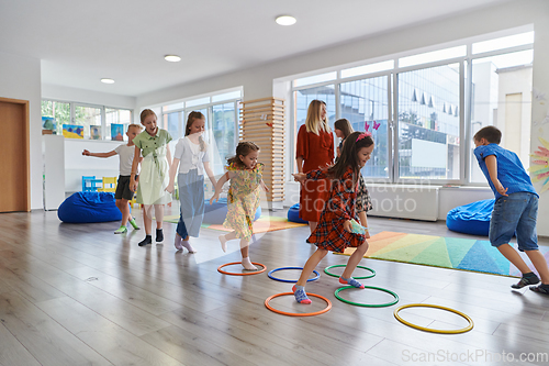 Image of Small nursery school children with female teacher on floor indoors in classroom, doing exercise. Jumping over hula hoop circles track on the floor.