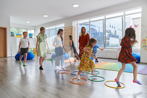 Image of Small nursery school children with female teacher on floor indoors in classroom, doing exercise. Jumping over hula hoop circles track on the floor.