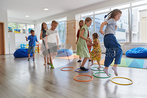 Image of Small nursery school children with female teacher on floor indoors in classroom, doing exercise. Jumping over hula hoop circles track on the floor.