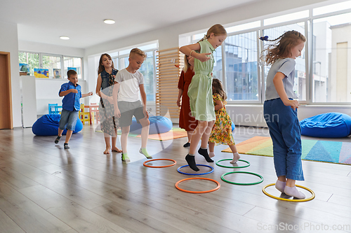 Image of Small nursery school children with female teacher on floor indoors in classroom, doing exercise. Jumping over hula hoop circles track on the floor.