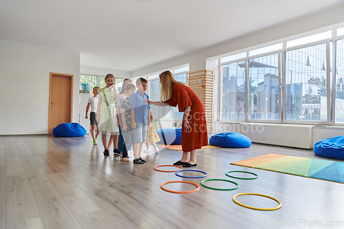 Image of Small nursery school children with female teacher on floor indoors in classroom, doing exercise. Jumping over hula hoop circles track on the floor.