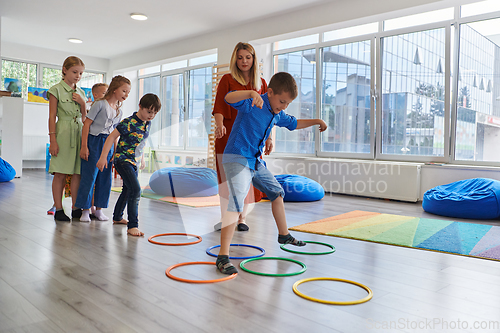 Image of Small nursery school children with female teacher on floor indoors in classroom, doing exercise. Jumping over hula hoop circles track on the floor.