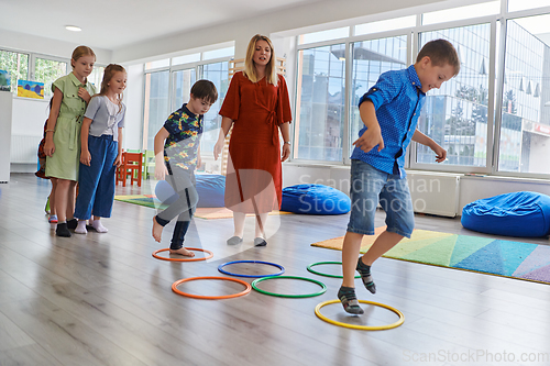 Image of Small nursery school children with female teacher on floor indoors in classroom, doing exercise. Jumping over hula hoop circles track on the floor.