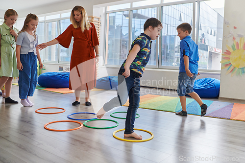 Image of Small nursery school children with female teacher on floor indoors in classroom, doing exercise. Jumping over hula hoop circles track on the floor.