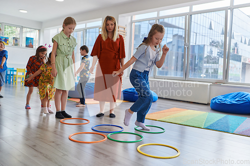 Image of Small nursery school children with female teacher on floor indoors in classroom, doing exercise. Jumping over hula hoop circles track on the floor.