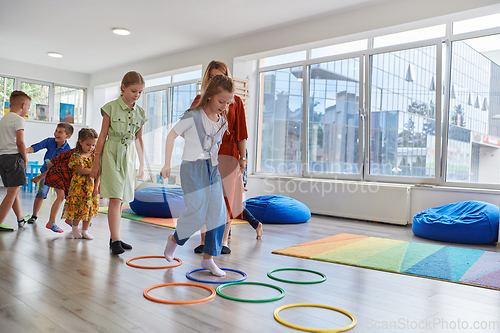 Image of Small nursery school children with female teacher on floor indoors in classroom, doing exercise. Jumping over hula hoop circles track on the floor.