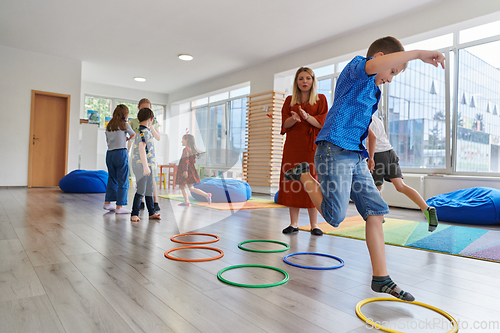 Image of Small nursery school children with female teacher on floor indoors in classroom, doing exercise. Jumping over hula hoop circles track on the floor.