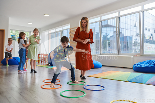 Image of Small nursery school children with female teacher on floor indoors in classroom, doing exercise. Jumping over hula hoop circles track on the floor.