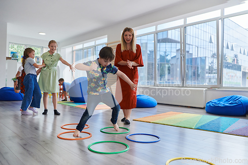 Image of Small nursery school children with female teacher on floor indoors in classroom, doing exercise. Jumping over hula hoop circles track on the floor.