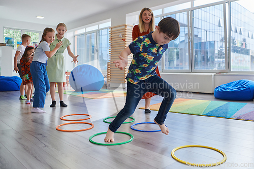 Image of Small nursery school children with female teacher on floor indoors in classroom, doing exercise. Jumping over hula hoop circles track on the floor.