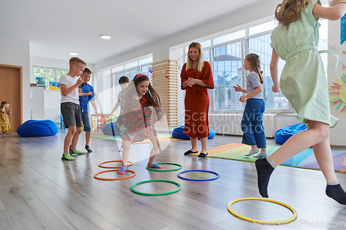 Image of Small nursery school children with female teacher on floor indoors in classroom, doing exercise. Jumping over hula hoop circles track on the floor.