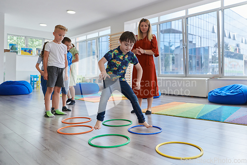 Image of Small nursery school children with female teacher on floor indoors in classroom, doing exercise. Jumping over hula hoop circles track on the floor.