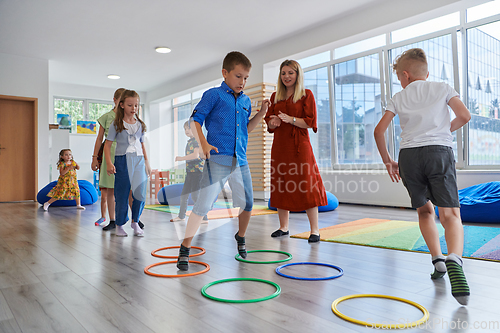 Image of Small nursery school children with female teacher on floor indoors in classroom, doing exercise. Jumping over hula hoop circles track on the floor.