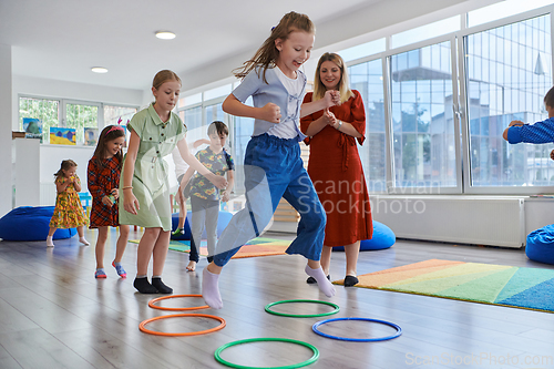 Image of Small nursery school children with female teacher on floor indoors in classroom, doing exercise. Jumping over hula hoop circles track on the floor.