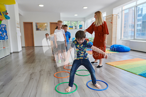 Image of Small nursery school children with female teacher on floor indoors in classroom, doing exercise. Jumping over hula hoop circles track on the floor.