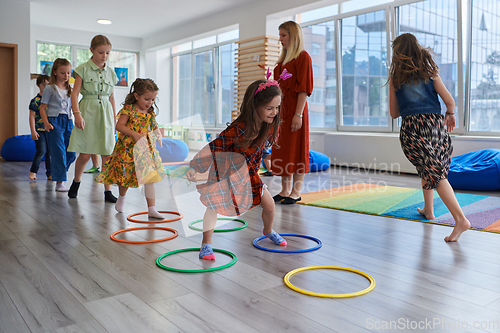 Image of Small nursery school children with female teacher on floor indoors in classroom, doing exercise. Jumping over hula hoop circles track on the floor.