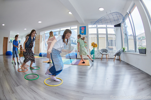 Image of Small nursery school children with female teacher on floor indoors in classroom, doing exercise. Jumping over hula hoop circles track on the floor.