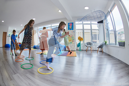 Image of Small nursery school children with female teacher on floor indoors in classroom, doing exercise. Jumping over hula hoop circles track on the floor.