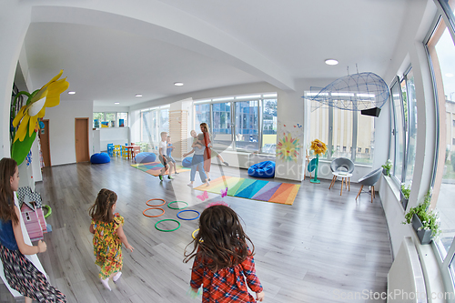 Image of Small nursery school children with female teacher on floor indoors in classroom, doing exercise. Jumping over hula hoop circles track on the floor.