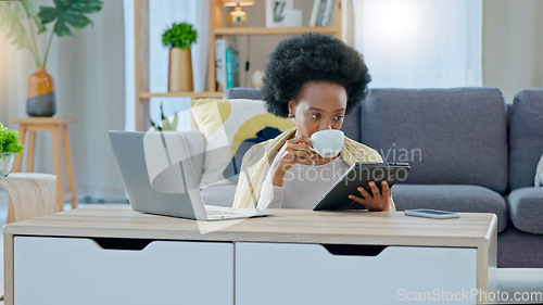 Image of African American woman working remotely sitting on the floor having coffee with her tablet in her hand , laptop on coffee table and her back against the sofa in a living room