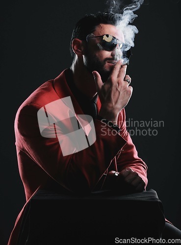 Image of Fashion, smoking and red with a man model in studio on a dark background wearing a suit for style. Smoke, mafia and edgy with a handsome young male posing on black space holding a cigarette