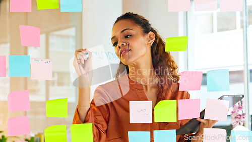 Image of A female designer planning ideas on a glass wall with colorful sticky notes inside a modern and creative office. Busy business woman enjoying her job while brainstorming projects and managing project