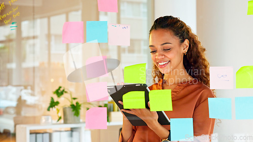 Image of Focused female designer planning ideas on a glass wall with colorful sticky notes inside a modern and creative office. Busy woman enjoying her job while brainstorming projects and managing projects