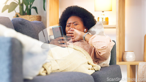 Image of Bored and tired woman lying on a couch typing on her phone at home. A young African female relaxing and yawing on a sofa in her house on a boring weekend. A sleepy lady scrolling social media