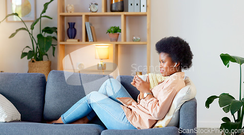 Image of Relaxed Black woman listening to a podcast while drinking coffee and sitting on a couch at home. Happy African American woman streaming online content, enjoying an audiobook while laughing