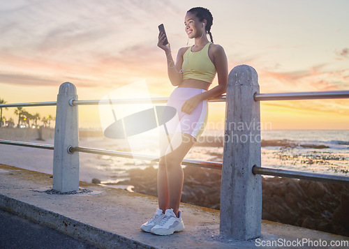 Image of Fitness, beach and woman with smartphone after running at sunset, online and streaming music or podcast in nature for exercise. Runner, relax and scroll social media after run, smile with earphones.