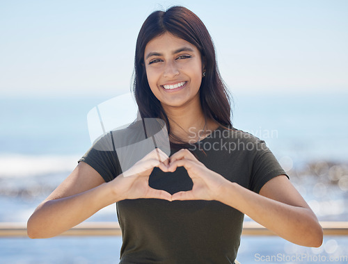 Image of Beach portrait and heart hand woman for summer holiday freedom, happiness and wellness. Happy Indian girl enjoying sun at ocean with love shape and optimistic smile for travel adventure.